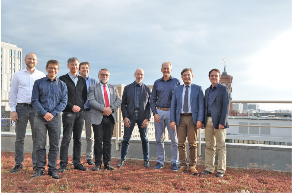 Zu sehen ist ein Gruppenfoto: 9 Männer in legeren Anzügen lächeln in die Kamera. Sie stehen auf einem Flachdach; im Hintergrund sind Hochhäuser und die Spitze des Roten Rathauses in Berlin zu sehen. 