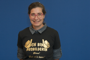 Zu sehen ist Andrea Kugler. Sie ist hat braune, hochgesteckte Haare, eine rote Brille und trägt ein schwarzes T-Shirt auf dem steht in goldener Schrift: "Ich bin Ausbilderin. Warum? Weil ich es kann!"