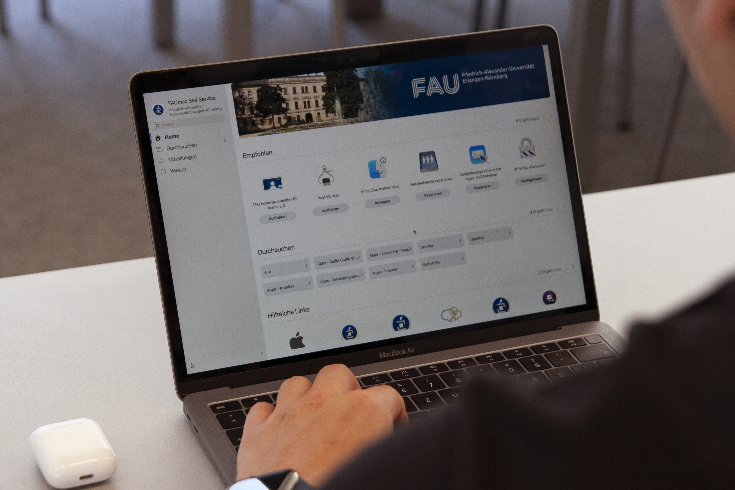 Ein Mann sitzt vor einem Mac und ruft den FAUmac-Self-Service auf. Zu sehen ist nur seine Schulter, die ein schwarzes T-Shirt kleidet und seine Hand mit einer Apple Watch. Neben dem PC liegt eine Schachtel mit AirPods.