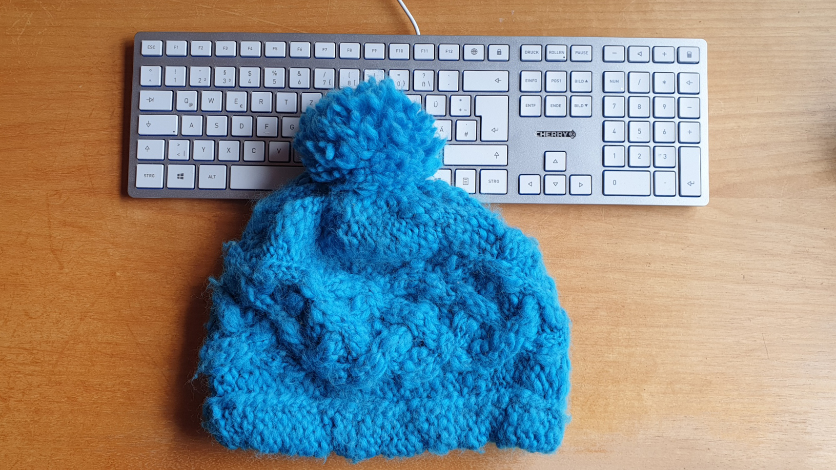 Zu sehen ist eine blaue Wollmütze. Sie liegt auf einer silber-weißen Tastatur auf einem Holztisch. 