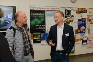 Zwei Männer im Gespräch: Der linke hat eine Glatze und trägt eine graue Jacke und ein kariertes Hemd. Der rechte Mann hat kurze Haare und einen Mehrtagebart. Er trägt eien schwarzen Anzug.