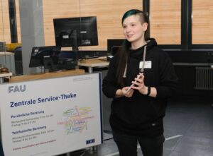 Eine Frau mit schwarz-grünen Haaren steht vor der Zentralen Service-Theke im RRZE, sie hält Moderationskarten in der Hand und trägt schwarze Kleidung.