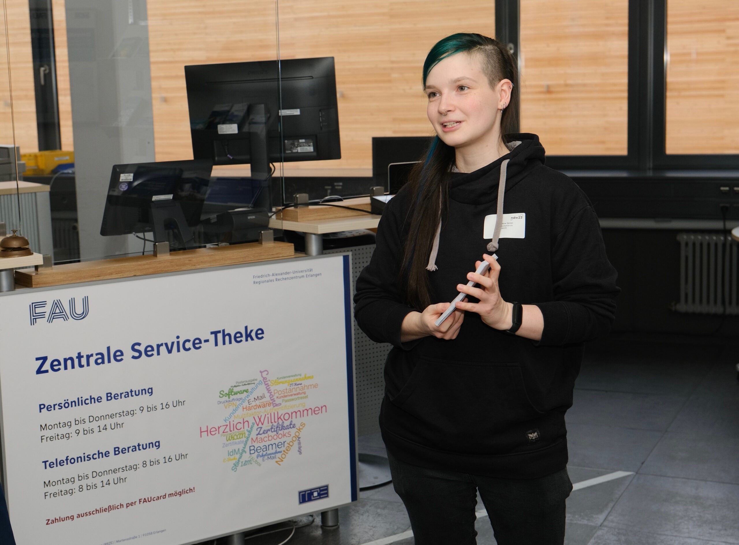 Eine Frau mit schwarz-grünen Haaren steht vor der Zentralen Service-Theke im RRZE, sie hält Moderationskarten in der Hand und trägt schwarze Kleidung.
