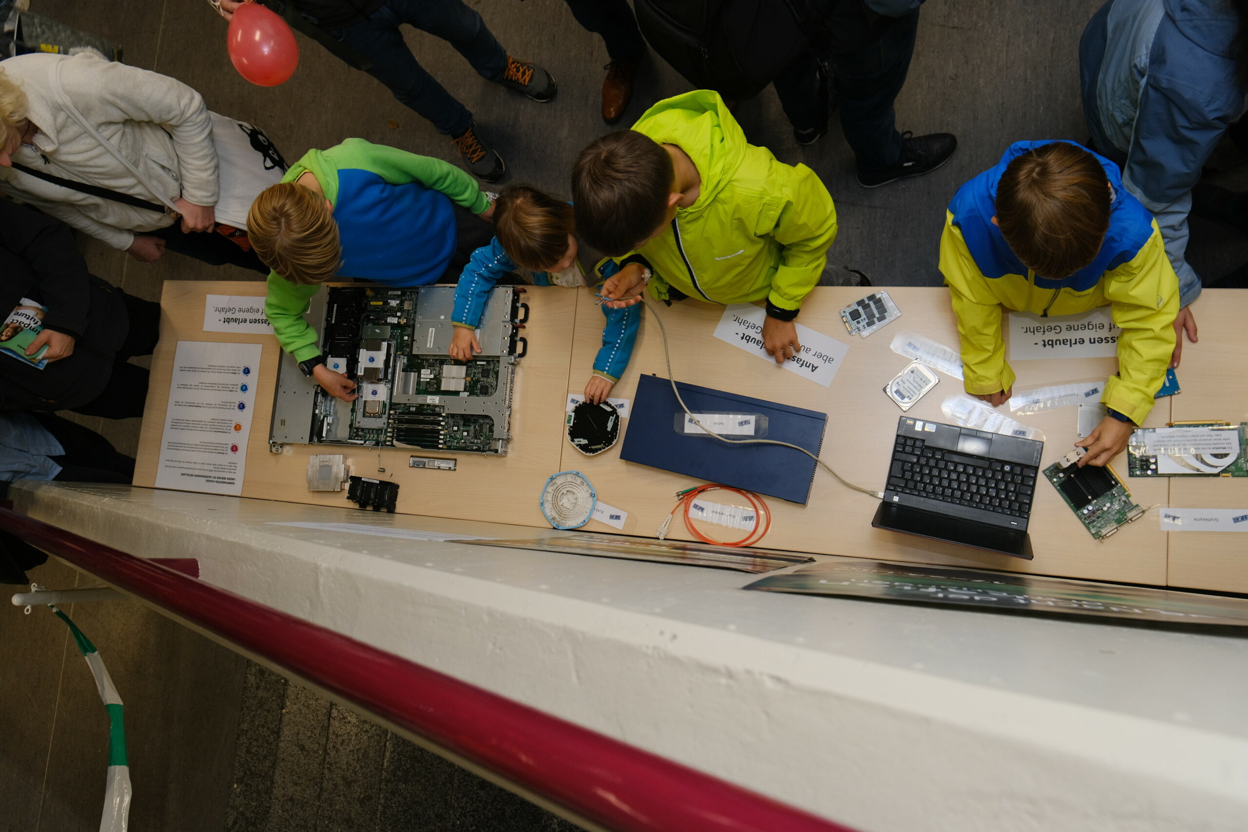 Blick von oben auf einen Tisch auf dem IT-Gegenstände liegen. Davor stehen drei Kinder in grün, blauen Jacken, die die Gegenstände genauer untersuchen.