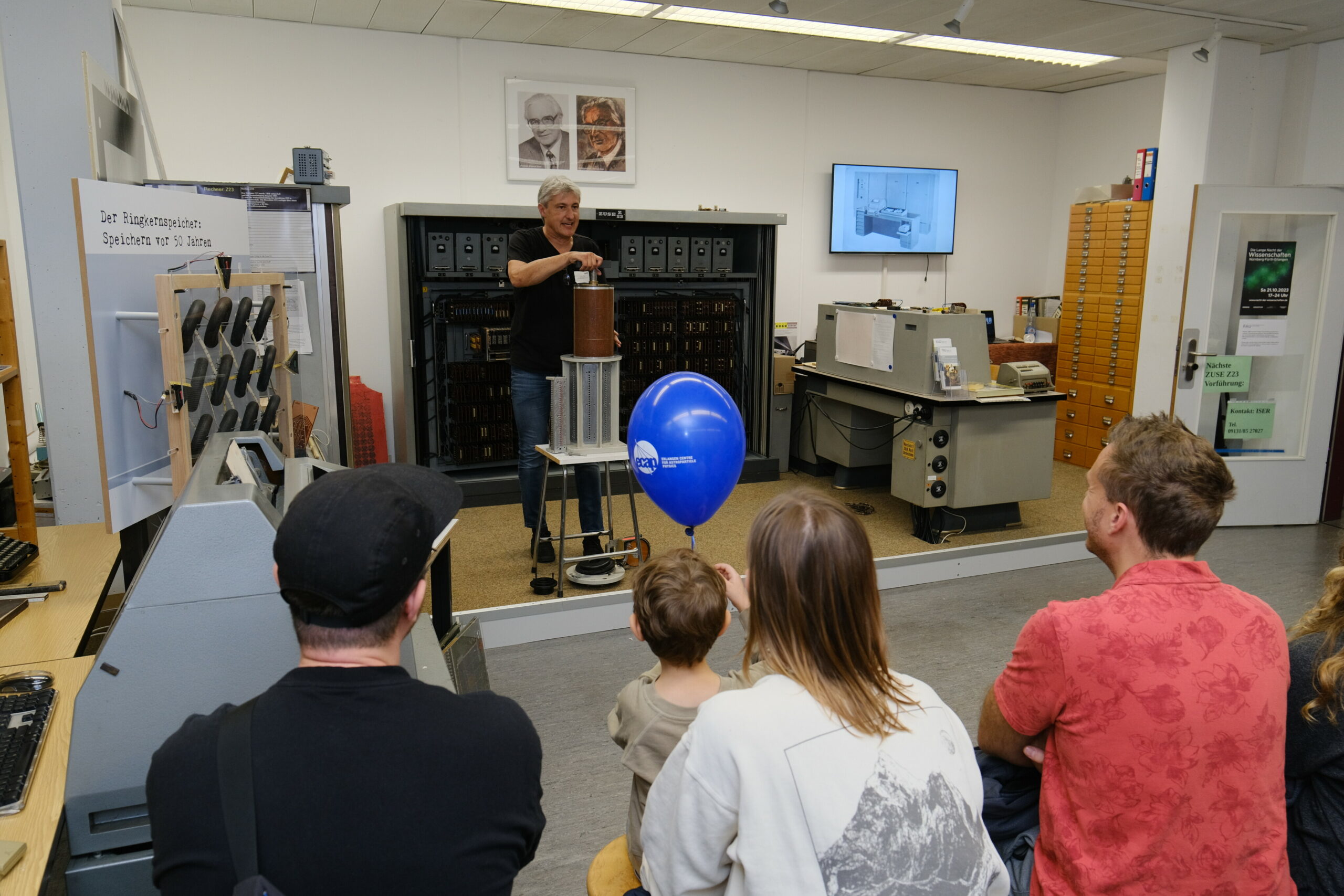 Im Hintergrund ist die ZUSE Z23 zu sehen, davor steht ein Mann mit grauen Haaren und erklärt diese. Die Zuschauer im Vordergrund hören interessiert zu.