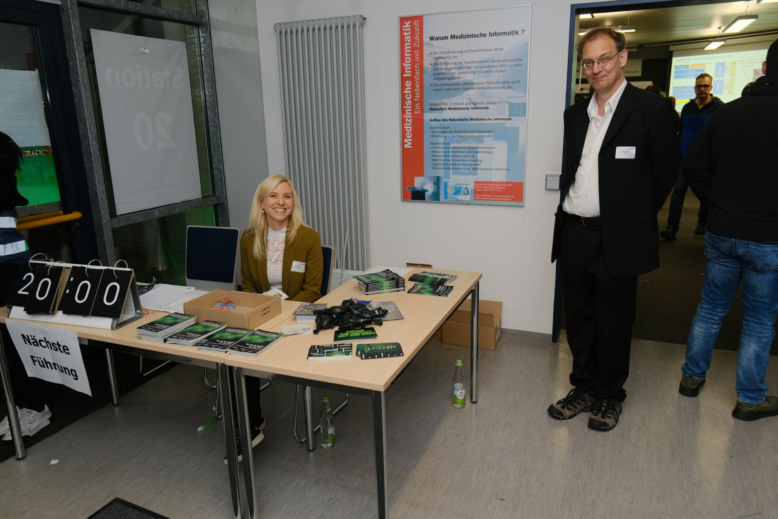 Im Foyer des RRZE stehen zwei Tische an denen eine blonde Frau sitzt. Daneben steht ein Mann mit kurzen Haaren und schwarzem Anzug.