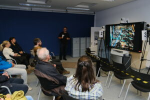 Besucher sitzen in einem Halbkreis vor einem Bildschirm auf dem der Serverraum des RRZE zu sehen ist. Im Hintergrund steht ein schwarz gekleideter Mann, der den Serverraum erklärt.
