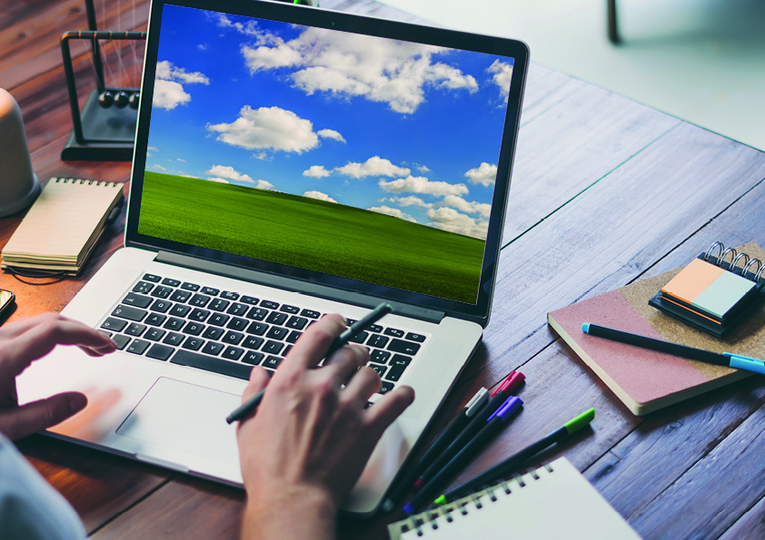 Ein Laptop steht auf einem Schreibtisch. Auf der Tastatur sind zwei Hände zu sehen. Der Bildschirm zeigt den typischen Windows Hügel, ein grün begraster Hügel unter blauem Himmel mit weißen Wolken.