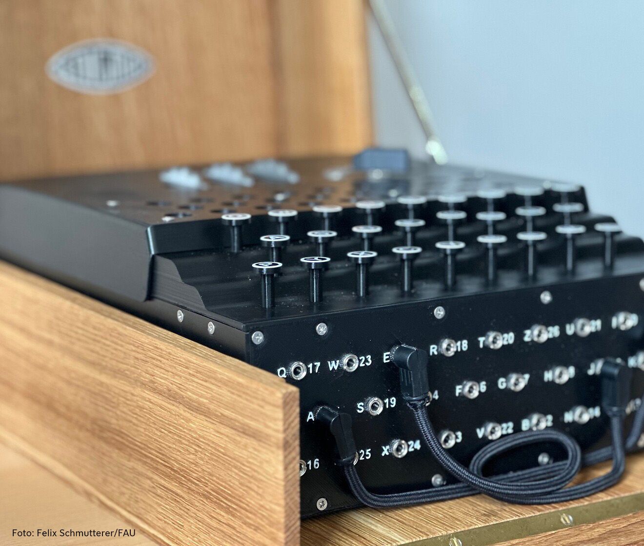 Ein Holzkasten mit einem Gerät, das Ähnlichkeiten mit einer Schreibmaschine aufweist. Oben sind Tasten mit Buchstaben, die man reindrücken kann. Drei Walzen sind oben und vorne können Steckerverbindungen gesetzt werden.