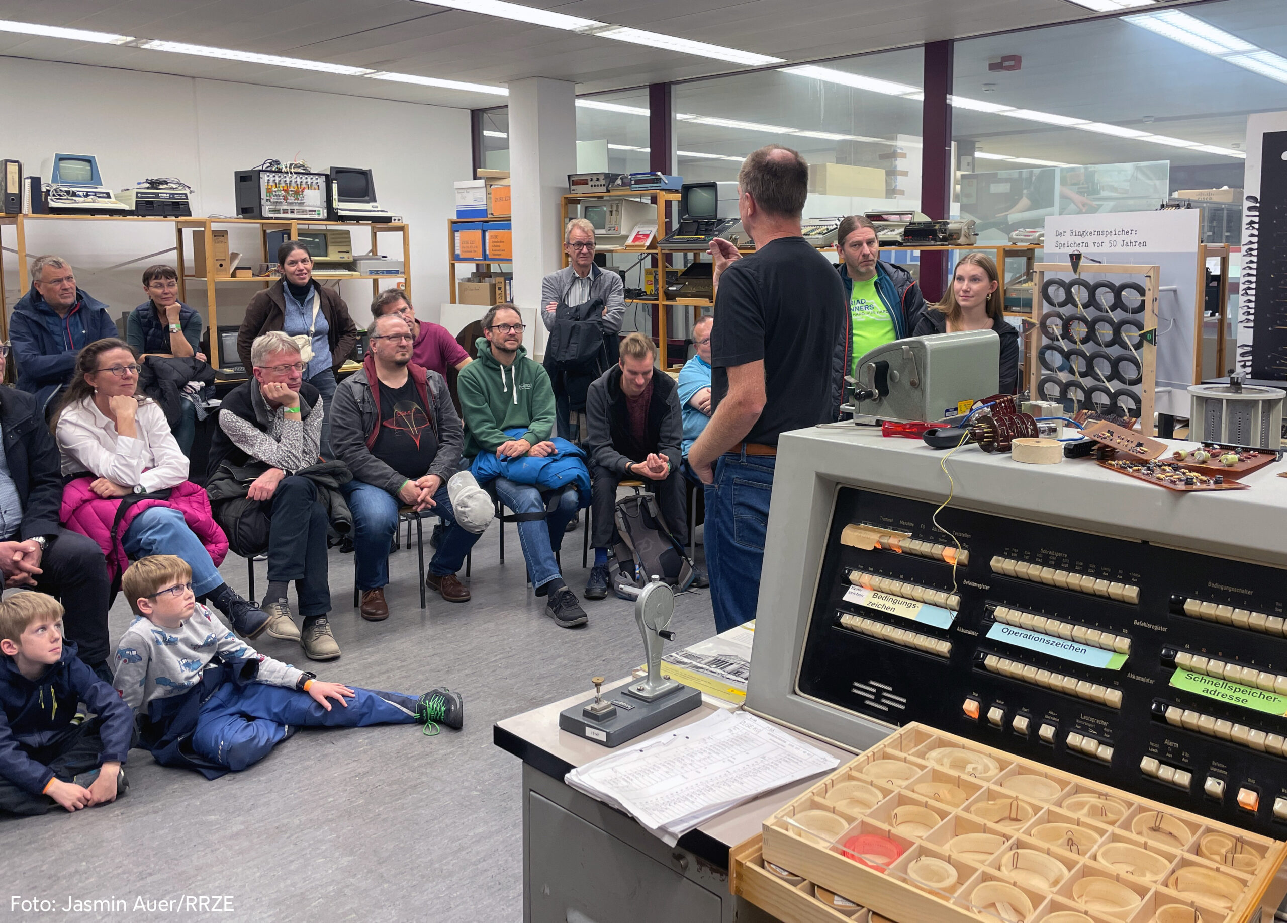 Im rechten Vordergrund steht die Steuerungskonsole einer Zuse Z23. Im Raum in einem Halbkreis auf Hockern sitzen Erwachsene. Vor ihnen am Boden sitzen zwei Jungen. Alle hören einem Mann in Jeans und schwarzem Oberteil zu.