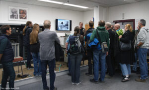 In einem historisch anmutenden Raum steht eine Menschenmenge und hört einem Mann zu, der auf einem Podest steht. Hinter den Menschen sieht man teilweise die Teile einer Zuse Z23.