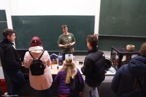 In einem Hörsaal von der ersten Sitzreihe aus fotografiert, steht vor der Tafel ein junger Mann, der lächelt und hält etwas in der Hand. Vor ihm stehen mit dem Rücken zur Kamera vier Personen, die sich mit ihm unterhalten. Auf dem Hörsaalpult steht ein 3D-Drucker und über den Tisch verteilt liegen Werkzeug, Filament und Druckbeispiele.
