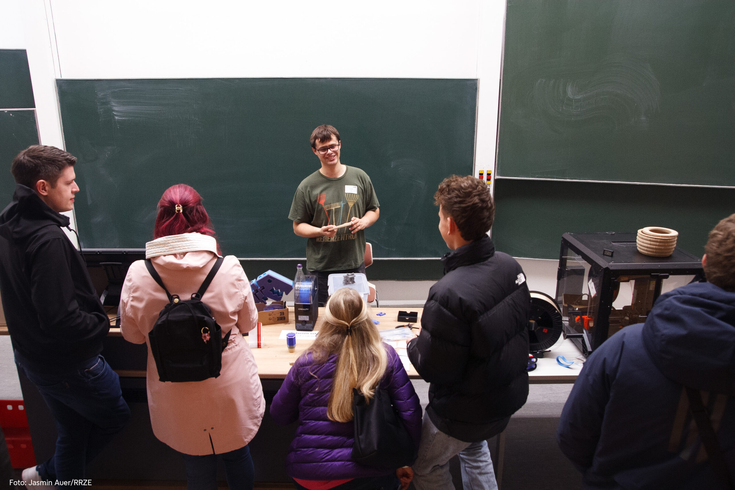 In einem Hörsaal von der ersten Sitzreihe aus fotografiert, steht vor der Tafel ein junger Mann, der lächelt und hält etwas in der Hand. Vor ihm stehen mit dem Rücken zur Kamera vier Personen, die sich mit ihm unterhalten. Auf dem Hörsaalpult steht ein 3D-Drucker und über den Tisch verteilt liegen Werkzeug, Filament und Druckbeispiele.