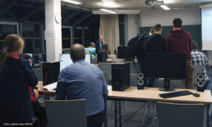 In einem großen Raum sitzen links im Vordergrund zwei erwachsene Menschen mit dem Rücken zur Kamera und schauen auf einen Bildschirm. Hinter ihnen steht ein Mädchen und schaut ihnen zu. Weiter vorne im Raum steht eine Gruppe junger Männer, der linke trägt einen schwarzen Rucksack und wird halb vom zweiten verdeckt, der zweite ist schwarz gekleidet und der dritte trägt einen dunkelroten Pulli und eine beige Hose. Ganz vorne im Raum sitzt noch eine Gruppe von vier Menschen.