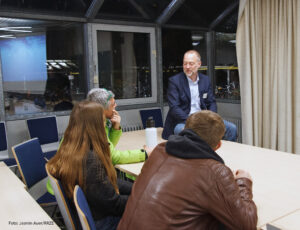 Ein Mann in blauem Sakko und hellblauem Hemd sitzt auf dem Tisch und unterhält sich mit drei weiteren Personen, links ein Mann mit grüner Jacke, stützt seinen Kopf auf seinen Arm, daneben eine Frau mit braunem, langem Haar und rechts ein Mann in brauner Lederjacke, die vor einem anderen Tisch sitzen. Im Hintergrund scheint die Nacht durch die Fenster.