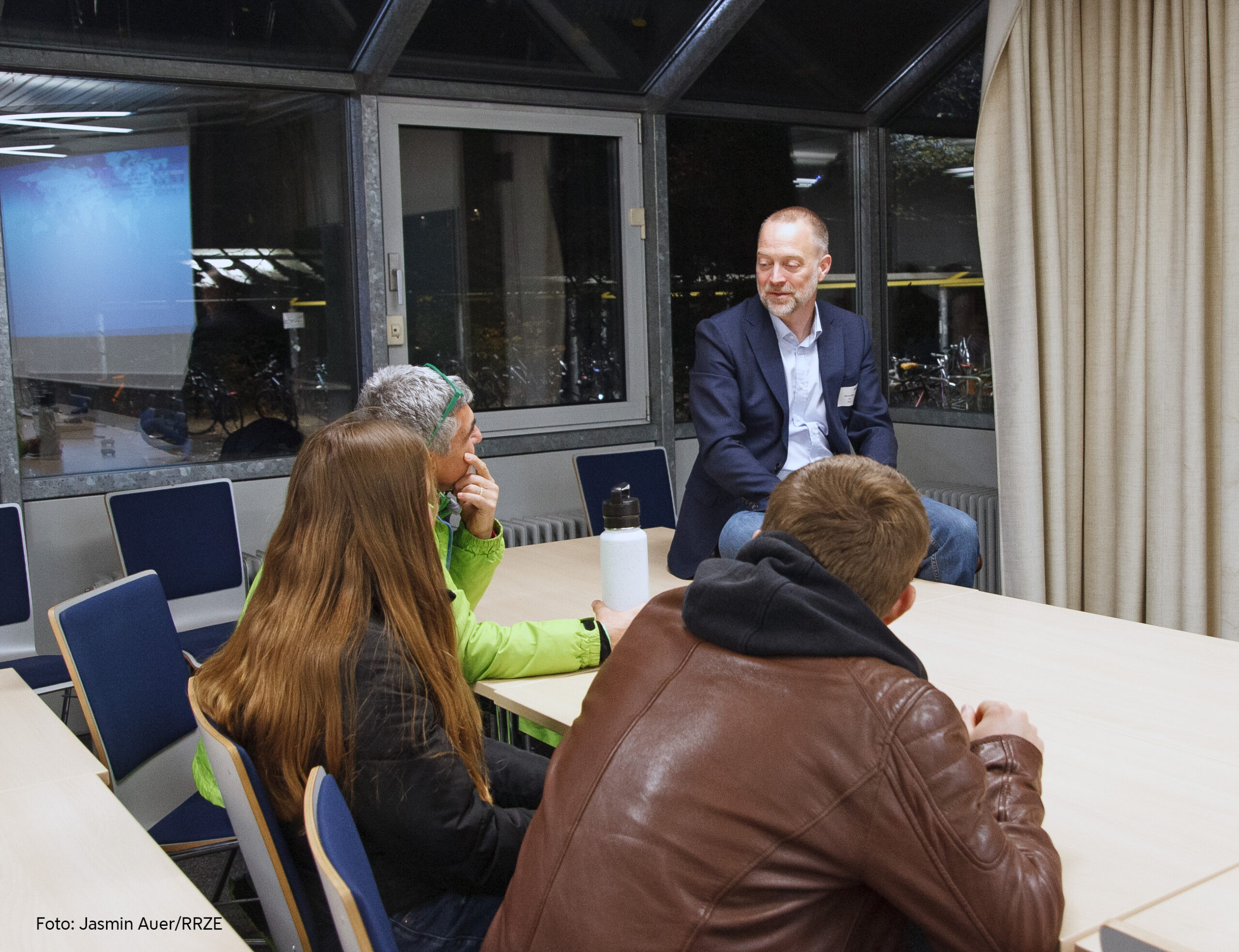 Ein Mann in blauem Sakko und hellblauem Hemd sitzt auf dem Tisch und unterhält sich mit drei weiteren Personen, links ein Mann mit grüner Jacke, stützt seinen Kopf auf seinen Arm, daneben eine Frau mit braunem, langem Haar und rechts ein Mann in brauner Lederjacke, die vor einem anderen Tisch sitzen. Im Hintergrund scheint die Nacht durch die Fenster.