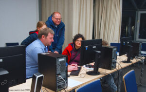 Zwei Personen sitzen vor einem Computer, zwei stehen hinter den Personen und schauen zu.
