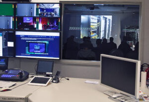 Im Vordergrund ist eine Regie zu sehen: Ein großer Tisch, auf dem mehrere Bildschirme stehen. Im Hintergrund ist ein Fenster in ein Studio. Dort sitzen im Dunkeln mehrere Personen, die nur schemenhaft zu sehen sind. Auf die Wand projiziiert wird ein Serverraum.