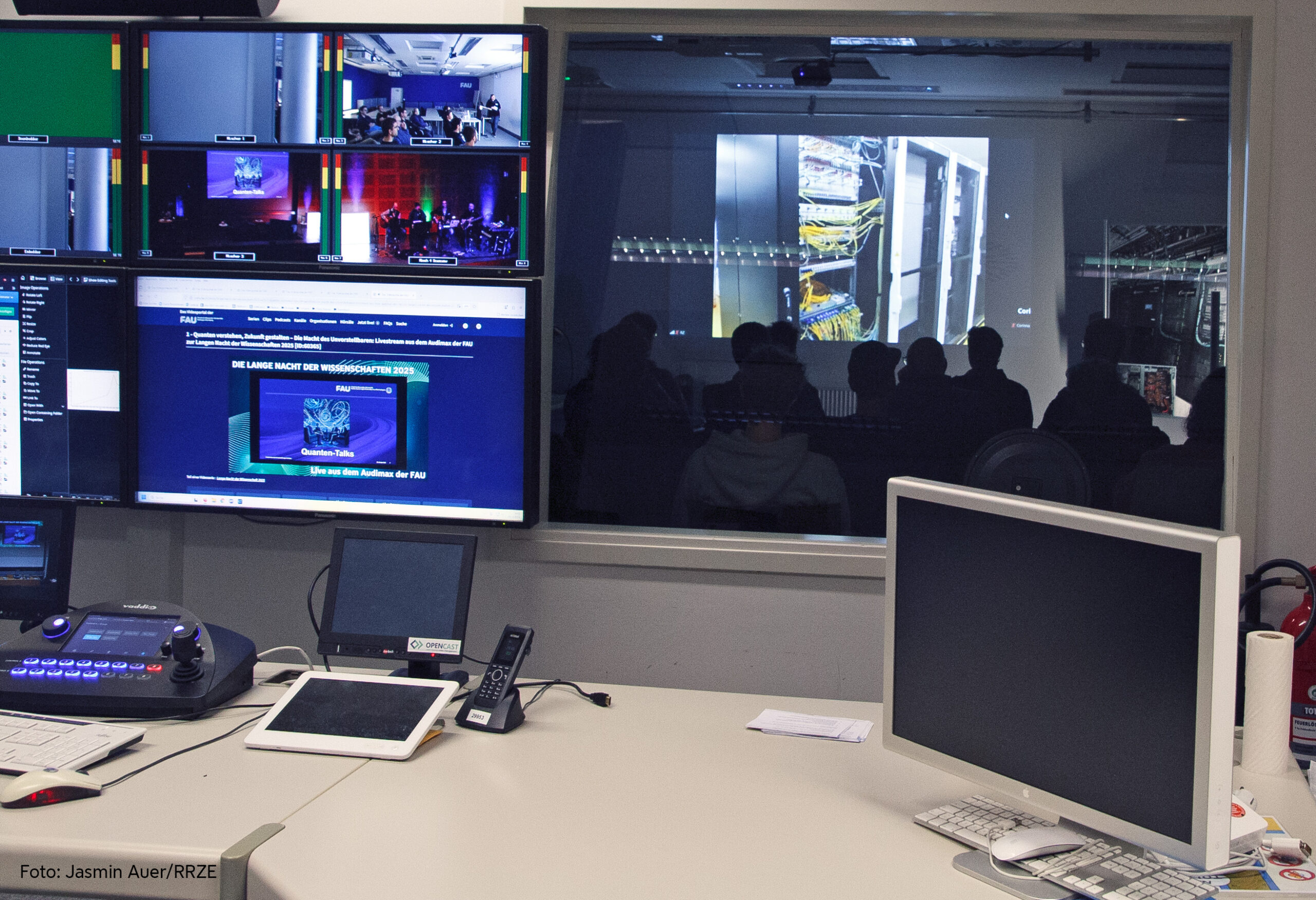 Im Vordergrund ist eine Regie zu sehen: Ein großer Tisch, auf dem mehrere Bildschirme stehen. Im Hintergrund ist ein Fenster in ein Studio. Dort sitzen im Dunkeln mehrere Personen, die nur schemenhaft zu sehen sind. Auf die Wand projiziiert wird ein Serverraum.