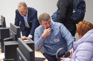 Im Vordergrund sitzen ein Mann und eine Frau vertieft in ein Gespräch. Der Mann trägt ein hellblaues Hemd, die Frau eien hellblaue Jacke. Auf dem Tisch vor ihnen stehen Computer inkl. Bildschirmen. Im Hintergrund steht eine weitere Person und beugt sich zu einem Rechner, um dort etwas einzustellen.