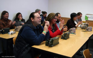 Im Fokus sitzen ein Mann und eine Frau, der Mann ist schwarz gekleidet, trägt langes Haar. Die Frau sitzt rechts daneben und trägt einen roten Pulli und eine Brille und offenes Haar. Vor ihnen auf dem Tisch stehen zwei Walther-Rechenmaschinen. Im Hintergrund sitzen weitere Menschen auch mit Rechenmaschinen auf dem Tisch.