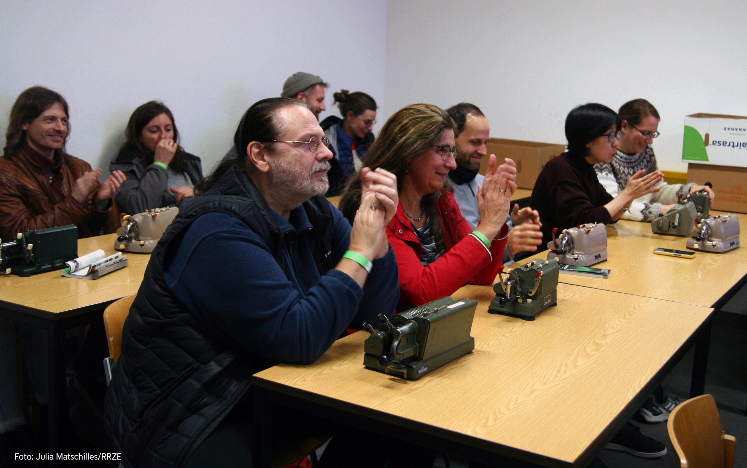 Im Fokus sitzen ein Mann und eine Frau, der Mann ist schwarz gekleidet, trägt langes Haar. Die Frau sitzt rechts daneben und trägt einen roten Pulli und eine Brille und offenes Haar. Vor ihnen auf dem Tisch stehen zwei Walther-Rechenmaschinen. Im Hintergrund sitzen weitere Menschen auch mit Rechenmaschinen auf dem Tisch.