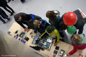 Von oben fotografierter Tisch, auf dem IT-Komponenten liegen. Davor stehen vier Personen, die die Komponenten teilweise in den Händen halten. Die rechte Person hält Luftballons in der Hand.