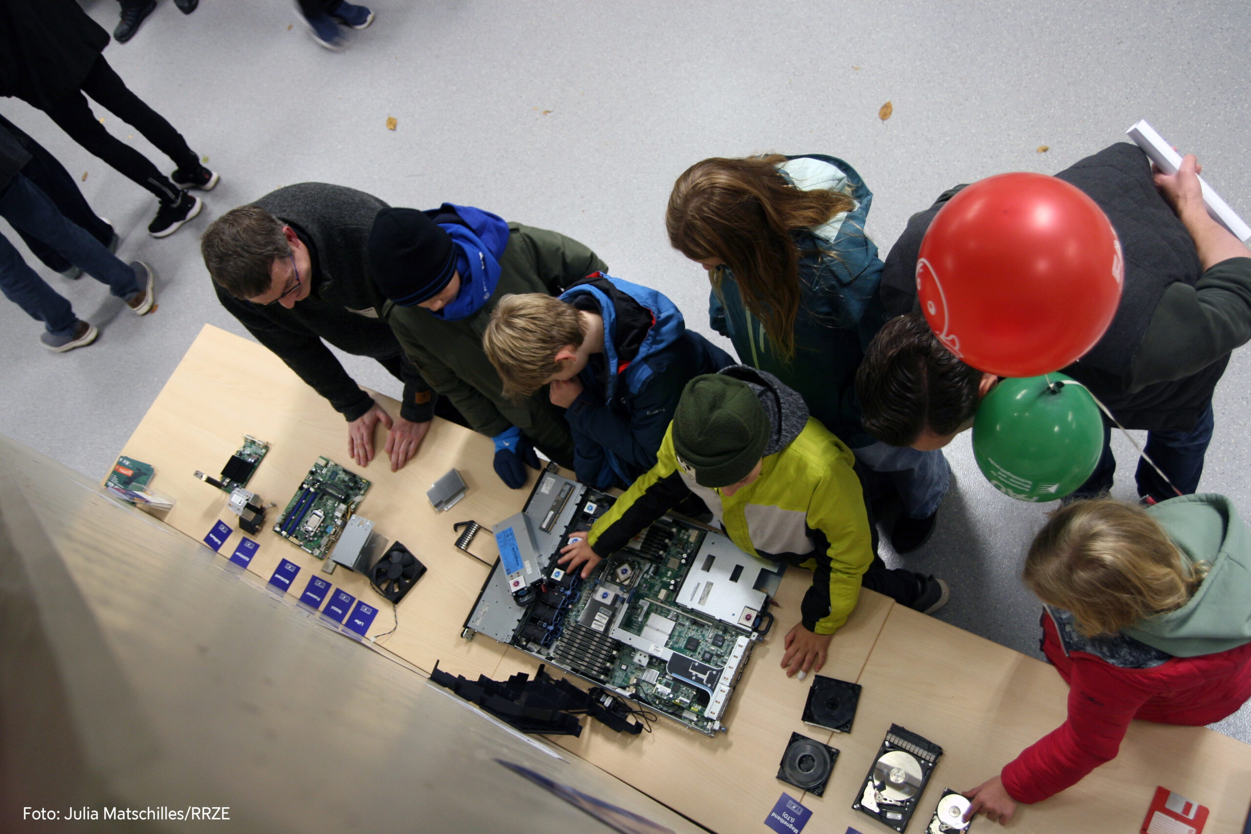 Von oben fotografierter Tisch, auf dem IT-Komponenten liegen. Davor stehen vier Personen, die die Komponenten teilweise in den Händen halten. Die rechte Person hält Luftballons in der Hand.